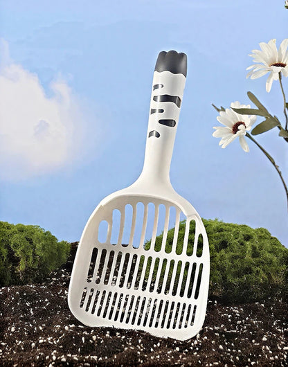 White cat litter scoop with black handle pattern in soil with green moss and white daisies background