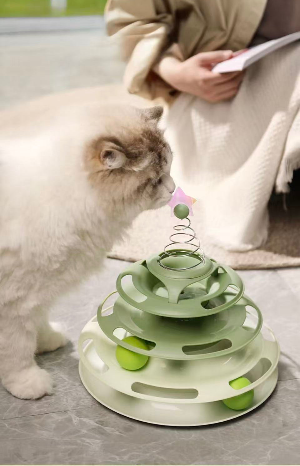 Fluffy white and gray cat playing with a green multi-tiered interactive ball toy indoors