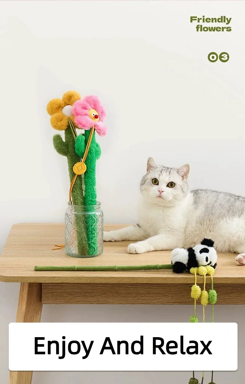 Gray and white cat lying on wooden table next to plush flower toys in glass jar and panda cat toy